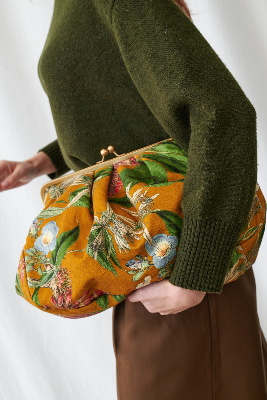One Hundred Stars Rhodoleia Gold Kiss Lock Clutch Bag. Person holding a floral clutch bag with a green sweater and brown skirt.