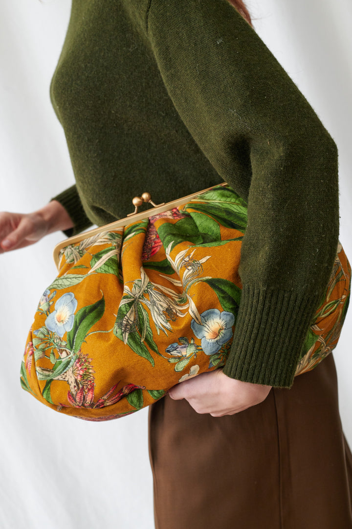 One Hundred Stars Rhodoleia Gold Kiss Lock Clutch Bag. Person holding a floral clutch bag with a green sweater and brown skirt.