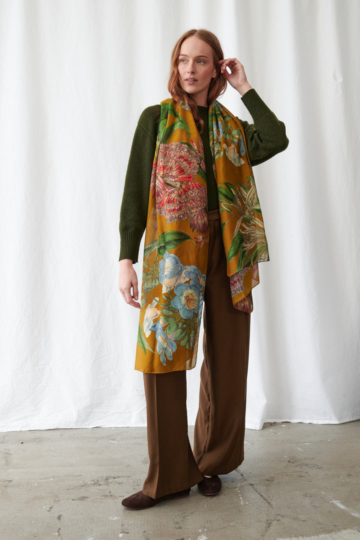 One Hundred Stars Rhodoleia Gold Scarf. Woman wearing a floral scarf with a green top and brown pants against a white background