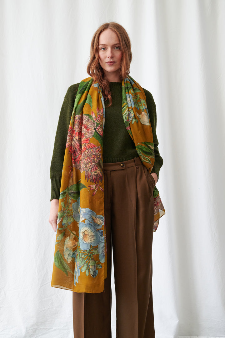 One Hundred Stars Rhodoleia Gold Scarf. Woman wearing a floral scarf with a green top and brown pants against a white background