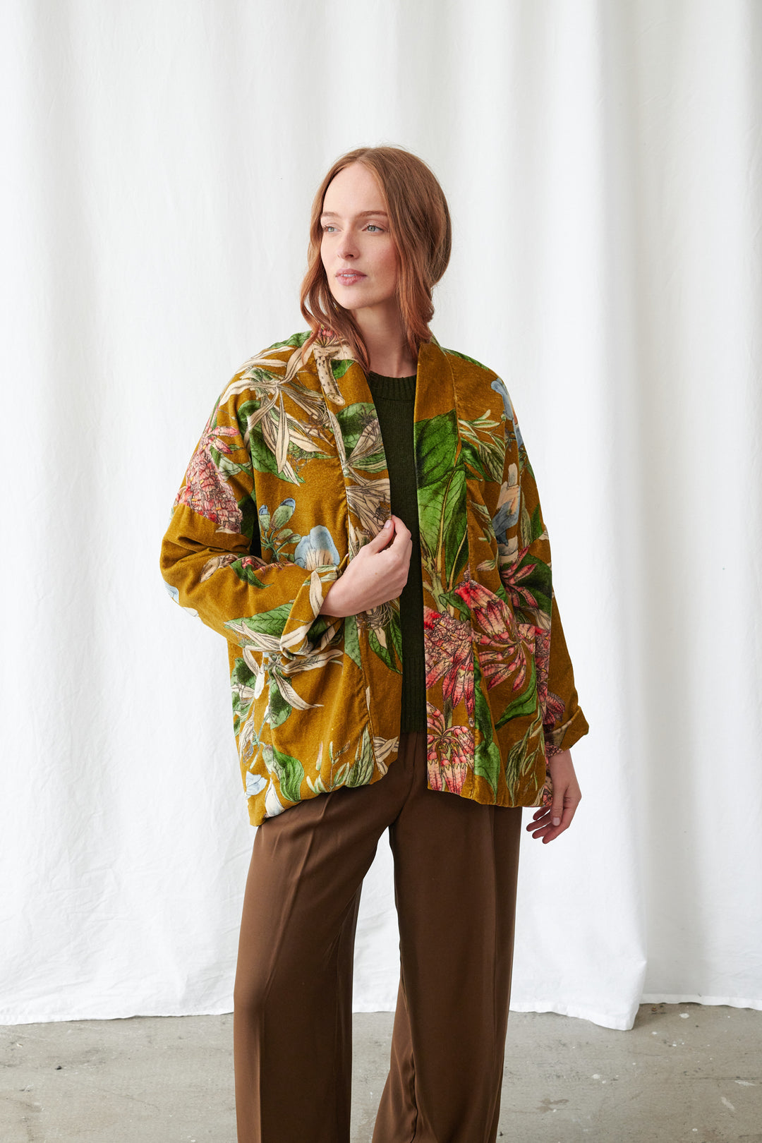 One Hundred Stars Rhodoleia Gold Velvet Pocket Jacket. Woman wearing a colorful floral jacket and brown pants against a white curtain background