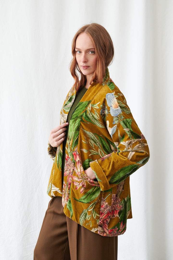 One Hundred Stars Rhodoleia Gold Velvet Pocket Jacket. Woman wearing a colorful floral jacket and brown pants against a white curtain background