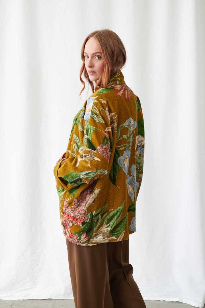 One Hundred Stars Rhodoleia Gold Velvet Pocket Jacket. Woman wearing a colorful floral jacket and brown pants against a white curtain background