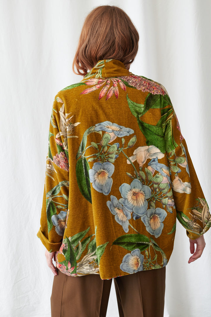 One Hundred Stars Rhodoleia Gold Velvet Pocket Jacket. Woman wearing a colorful floral jacket and brown pants against a white curtain background